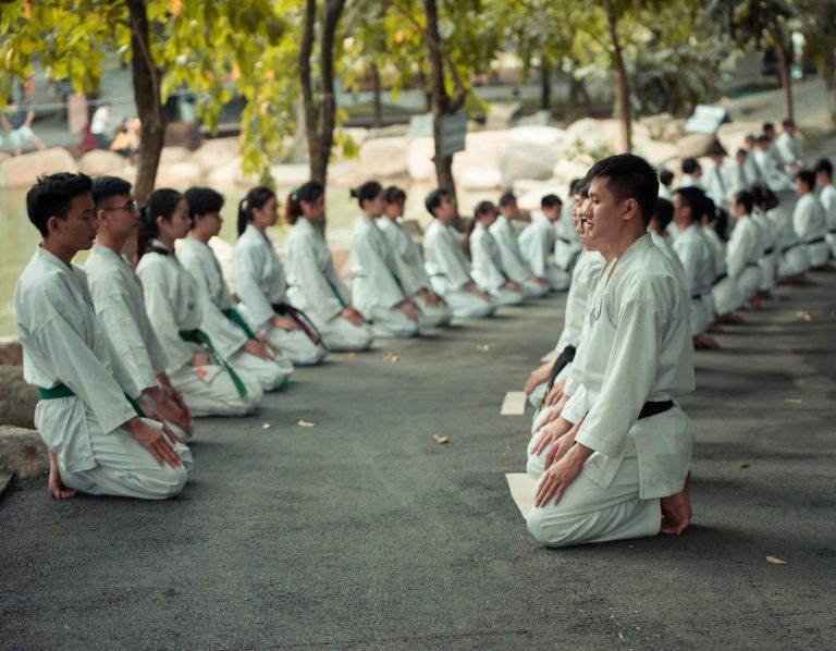 Taekwondo Gruppe in weißen Anzügen, sitzend und wartend in Reihen unter Bäumen.